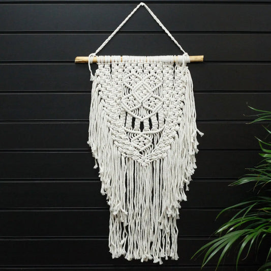Macramé decorativo blanco con flecos en forma de diamante, colgado en una barra de madera. Ideal para interiores.
