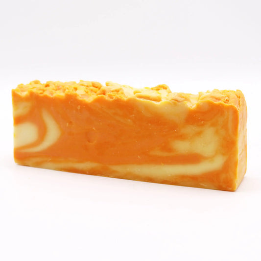 Barra de jabón artesanal rectangular con vetas en tonos naranja y crema, superficie superior rugosa sobre fondo blanco.