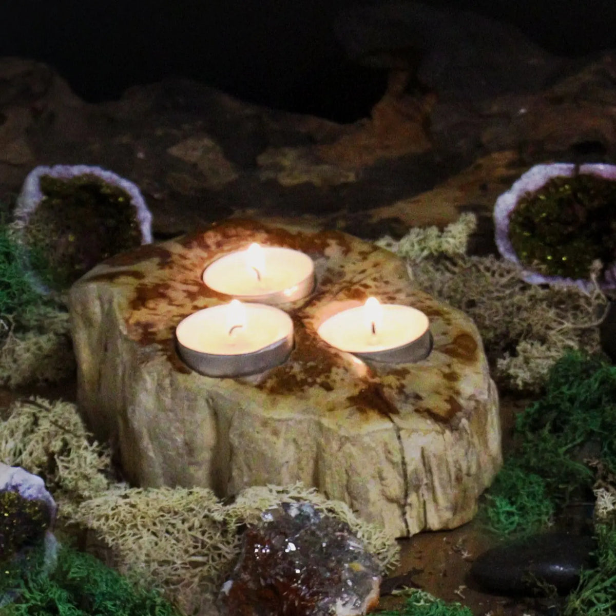 Portavelas de madera petrificada con tres velas encendidas. Textura rugosa y tonos tierra rodeado de musgo y cristales.