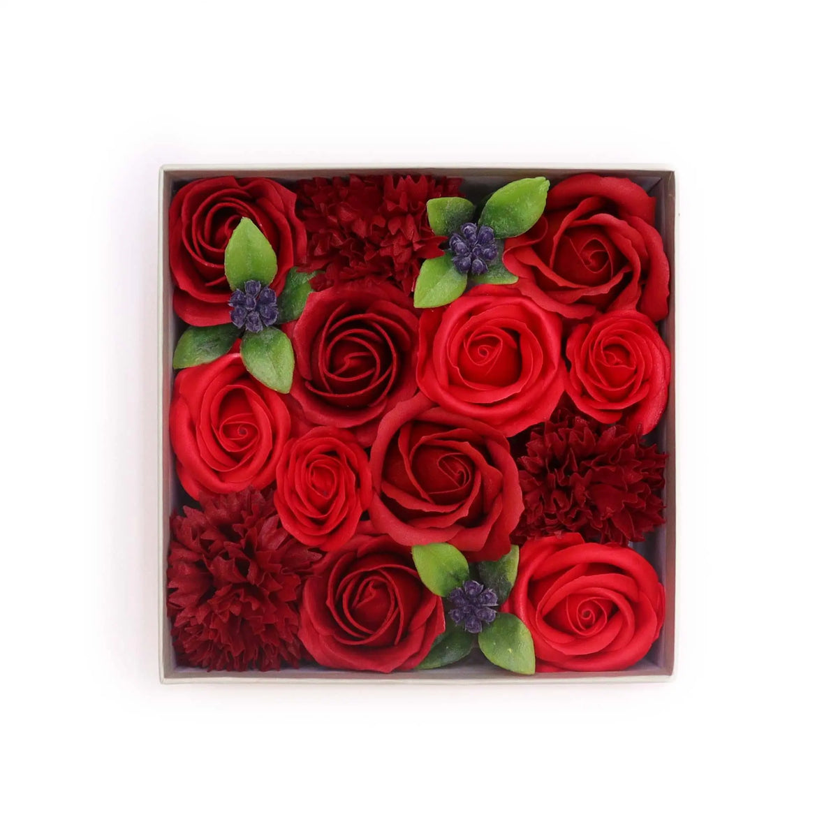 Caja cuadrada con rosas de jabón rojas, flores decorativas, hojas verdes y bayas moradas sobre fondo blanco.