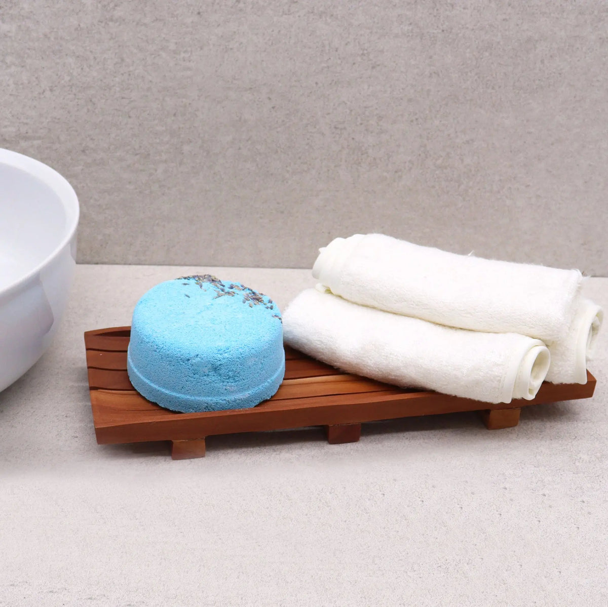 Bandeja de madera rectangular con barras, una pastilla de jabón azul y dos toallas blancas enrolladas sobre ella.