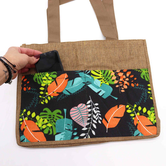 Bolsa de yute con asas beige y bolsillo exterior estampado con hojas tropicales coloridas sobre fondo negro.