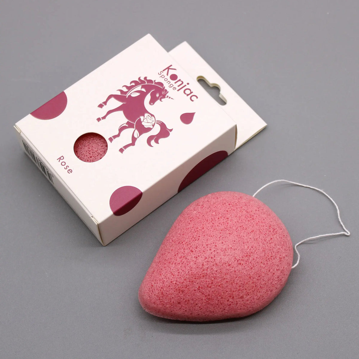 Esponja facial de konjac rosa en forma de gota con cordón junto a su caja de cartón con dibujo de unicornio.