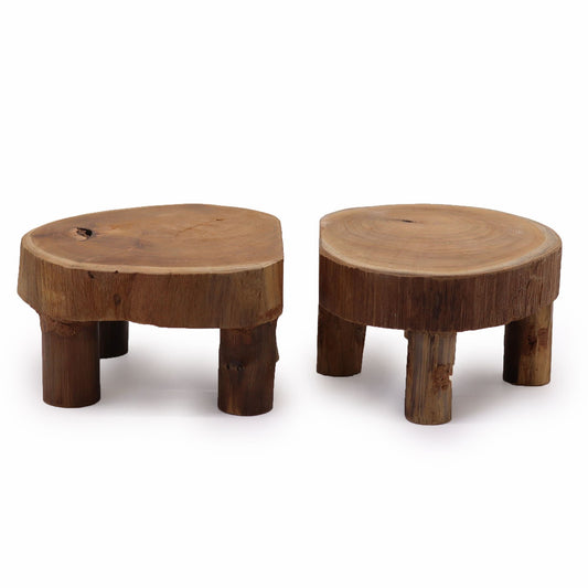 Dos soportes bajos de madera de teca natural con superficie redonda y rústica, sobre tres patas cortas.
