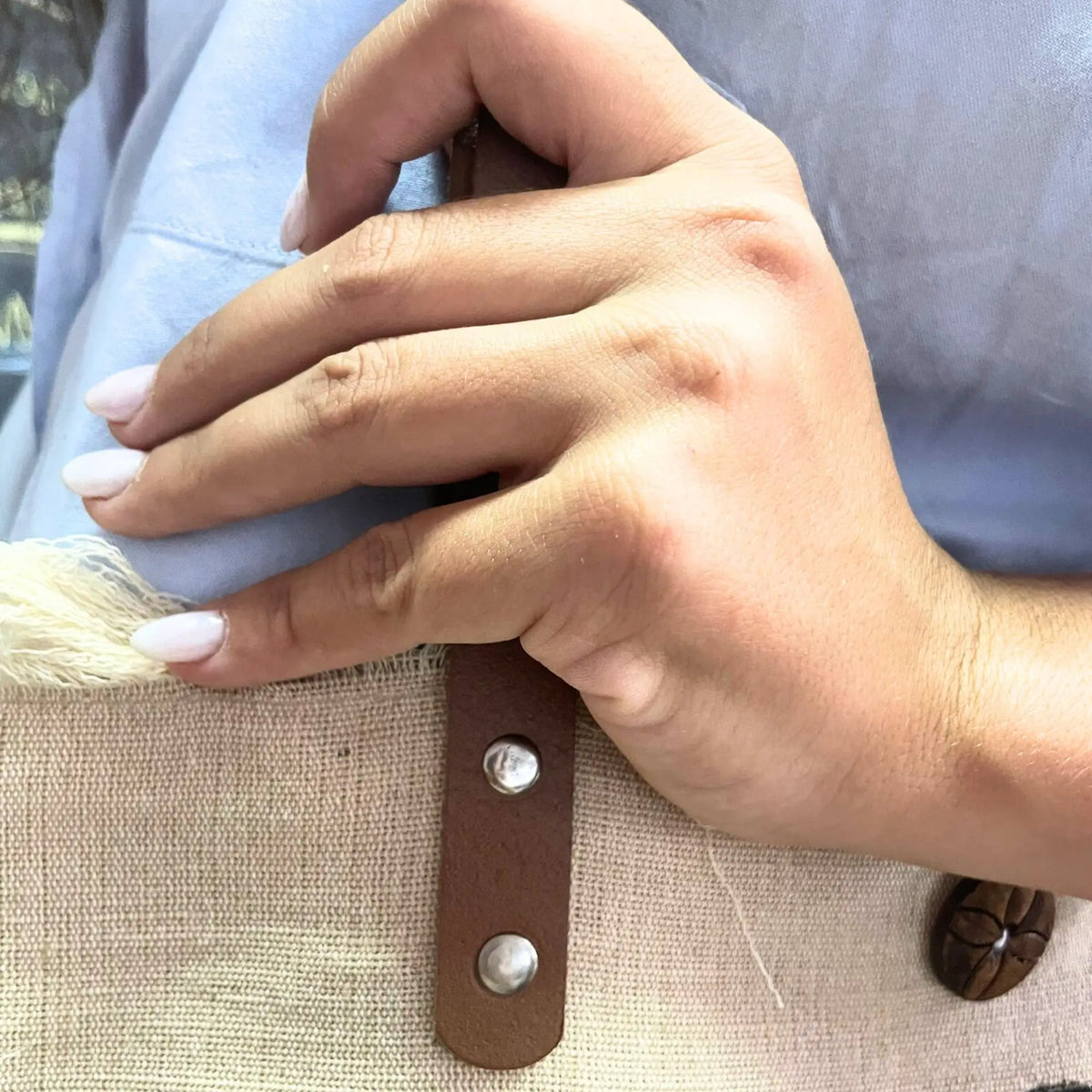 Primer plano de mano sujetando un bolso de tejido natural beige con asa de piel marrón fijada con remaches metálicos.