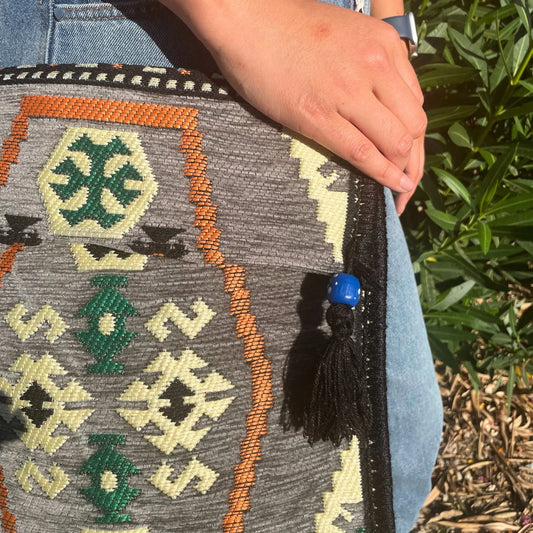 Bolso bandolera gris con tejido Kilim, motivos geométricos en naranja, verde y crema, y detalle de borla con cuenta azul.