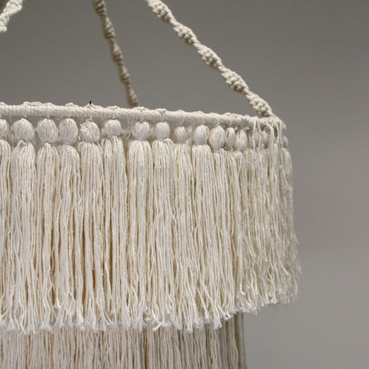 Lámpara colgante de macramé color crema, con diseño cilíndrico de dos niveles decorado con flecos largos y nudos.