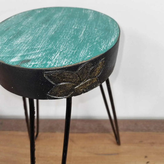 Soporte redondo de madera albasia turquesa desgastada, con borde negro y relieve floral dorado sobre patas de metal.