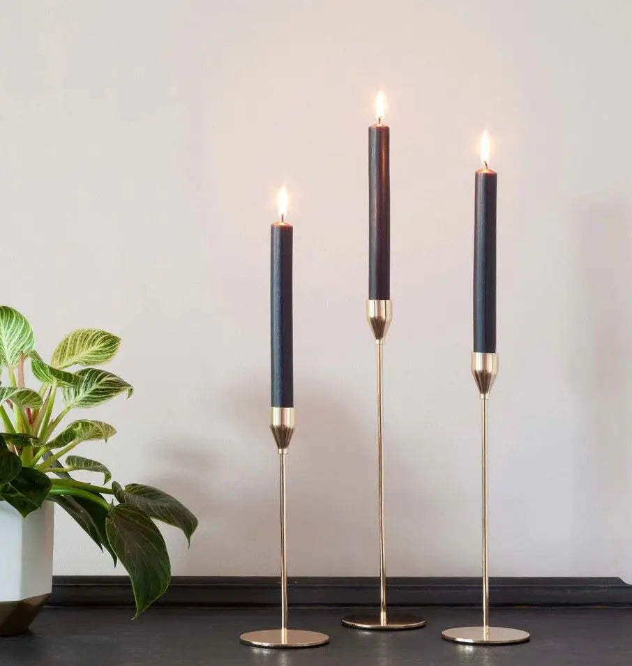 Tres velas negras rústicas encendidas sobre elegantes candelabros dorados de pie frente a una pared neutra y una planta
