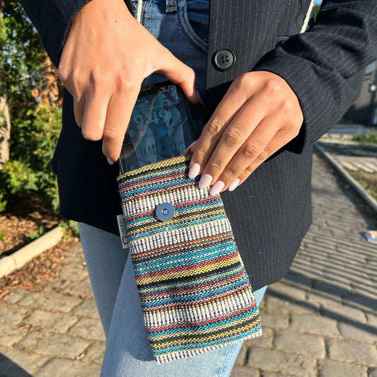 Bolso para móvil tejido a rayas multicolor con cierre de broche, sostenido por una persona con americana y jeans.