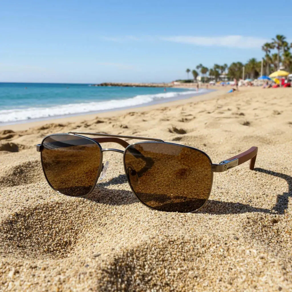 Gafas de sol tipo aviador con marco metálico, lentes marrones y patillas de madera sobre la arena de una playa.