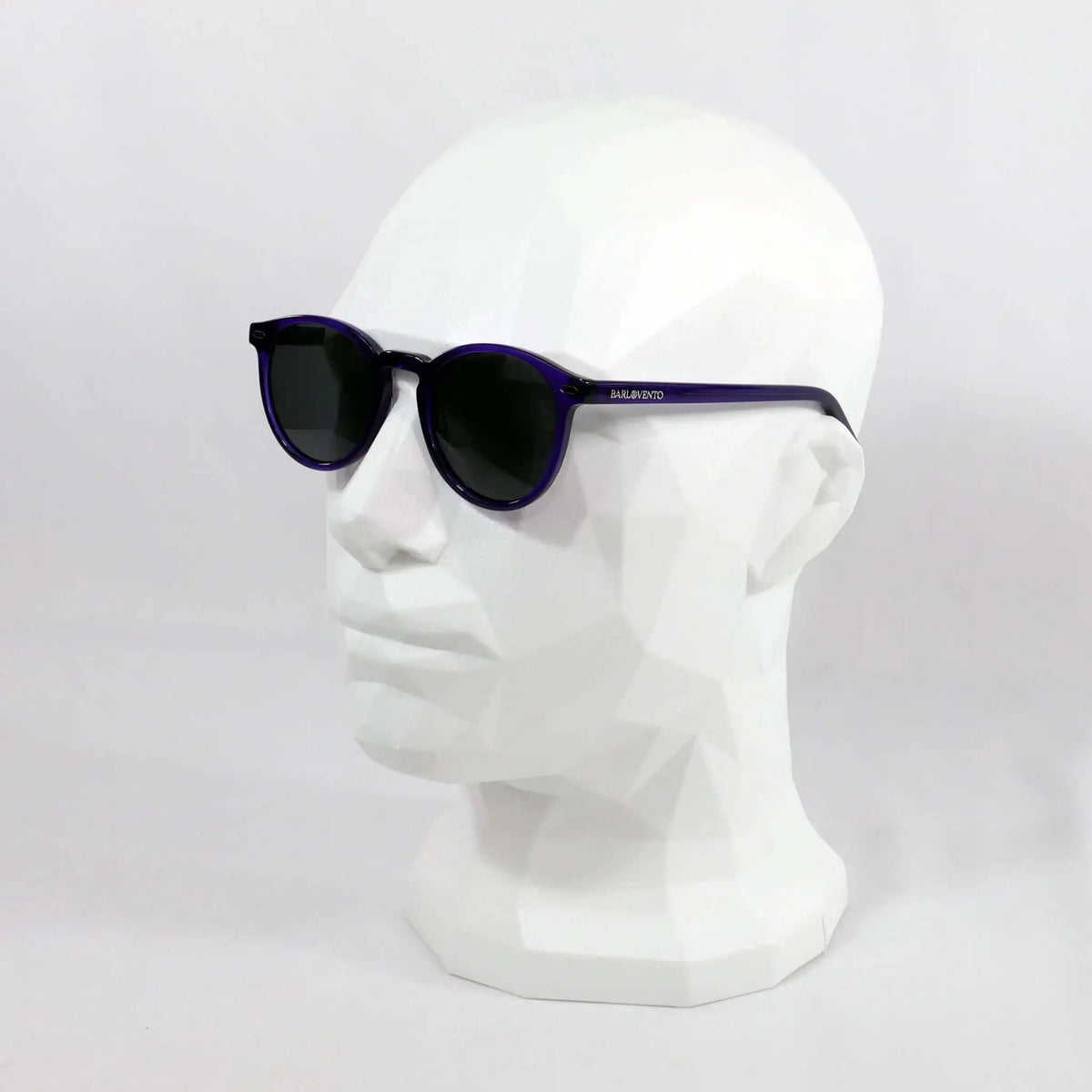 Gafas de sol de montura redonda en color morado traslúcido y cristales oscuros sobre un maniquí blanco.