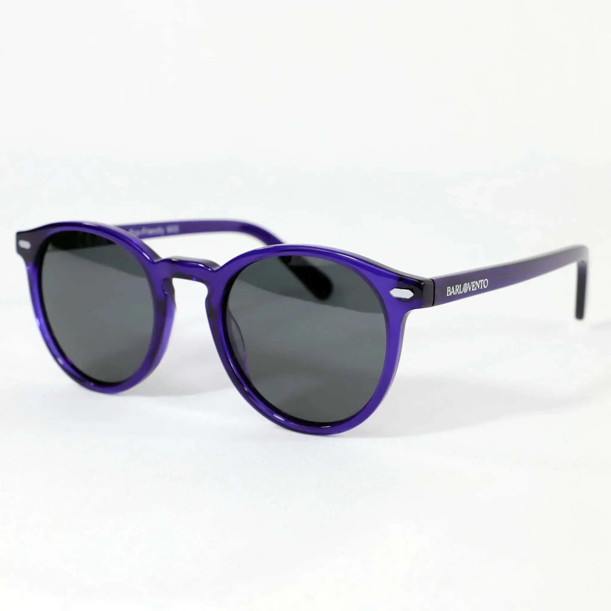 Gafas de sol redondas de montura púrpura traslúcida y cristales oscuros sobre fondo blanco. Estilo moderno y elegante.