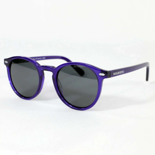 Gafas de sol redondas de montura púrpura traslúcida y cristales oscuros sobre fondo blanco. Estilo moderno y elegante.