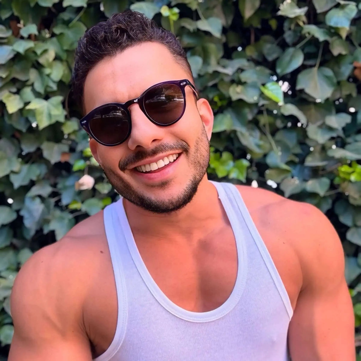 Hombre sonriente con gafas de sol redondas de montura púrpura oscura y cristales oscuros, estilo moderno y casual.