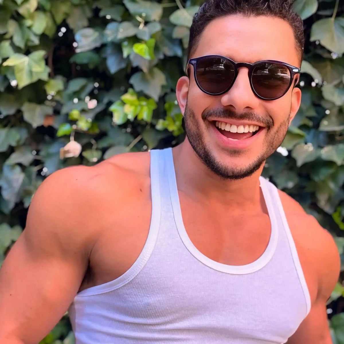 Hombre sonriente con gafas de sol redondas de montura oscura y cristales oscuros frente a un fondo de hojas verdes.