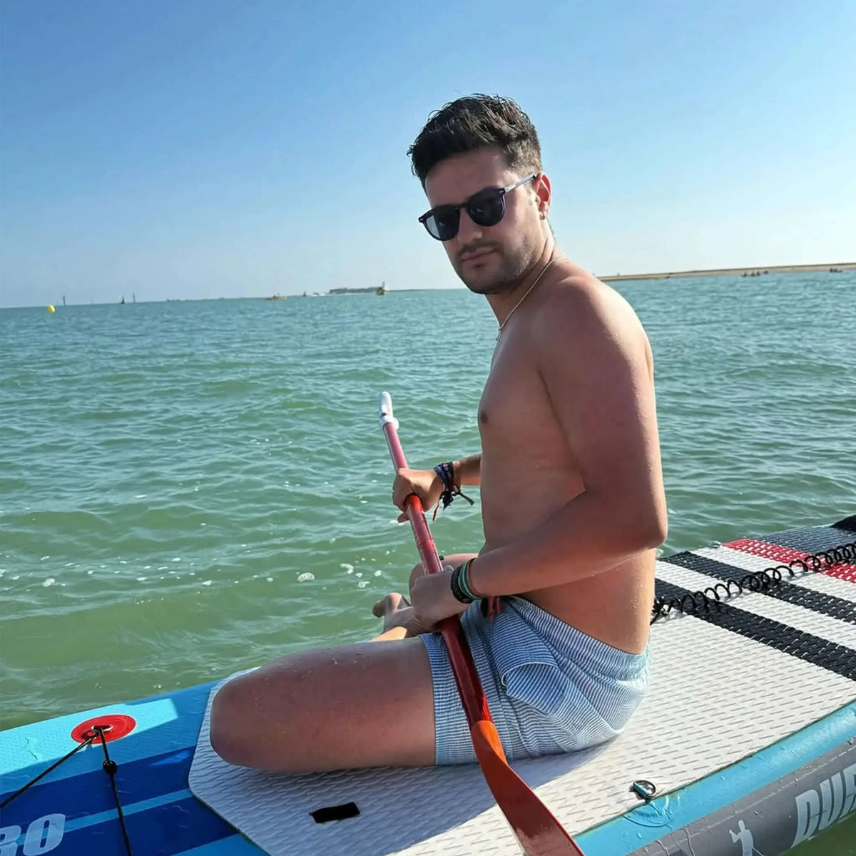 Hombre sentado sobre una tabla de surf con gafas de sol redondas de montura oscura y lentes oscuras.