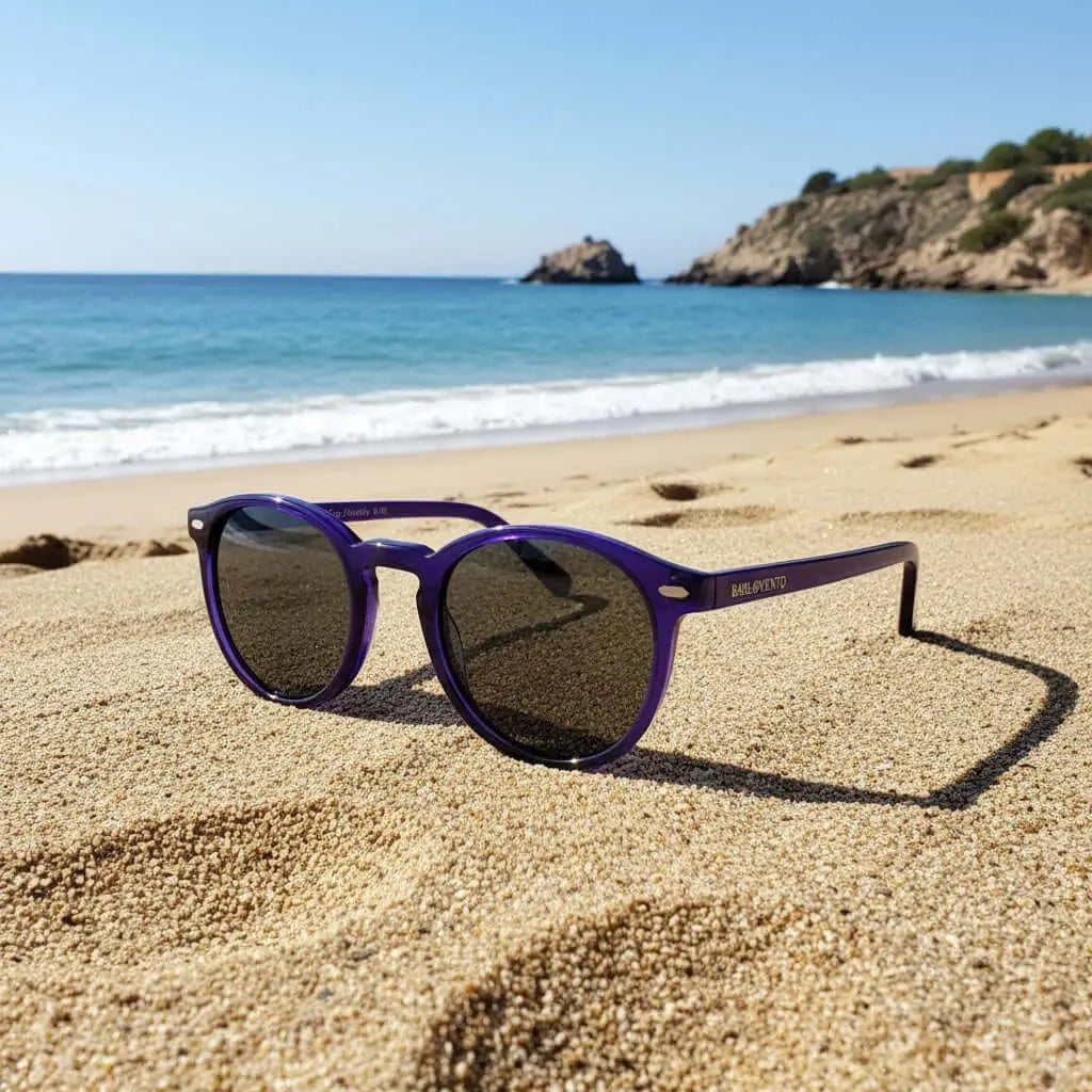 Gafas de sol redondas con montura de pasta color morado traslúcido y cristales oscuros sobre la arena de una playa.