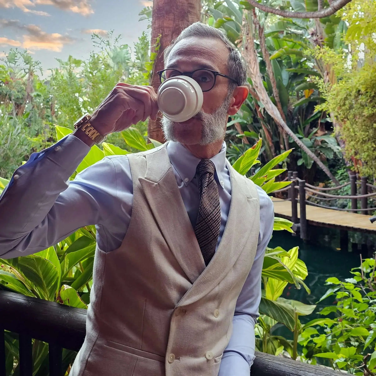 Hombre con camisa azul, chaleco beige y corbata de lana marrón con textura en un entorno tropical.