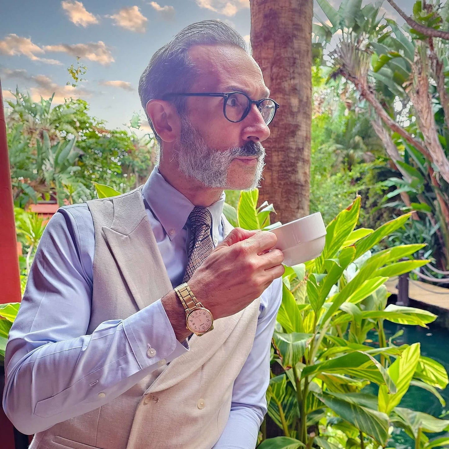 Hombre con barba, vestido elegante, corbata de lana marrón sosteniendo una taza de café, rodeado de vegetación exuberante.
