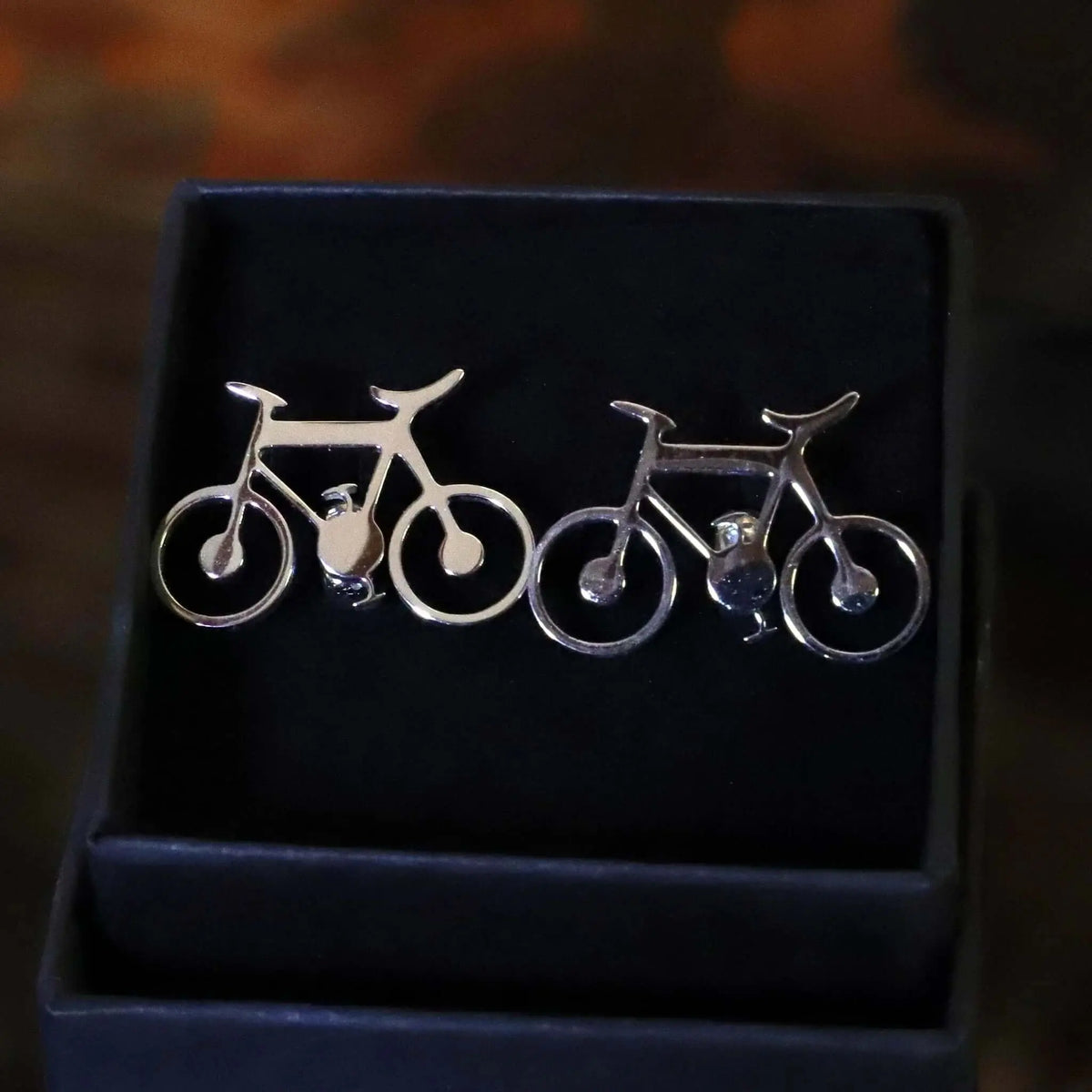 Gemelos plateados con forma de bicicleta minimalista, presentados en una caja de regalo oscura de terciopelo.