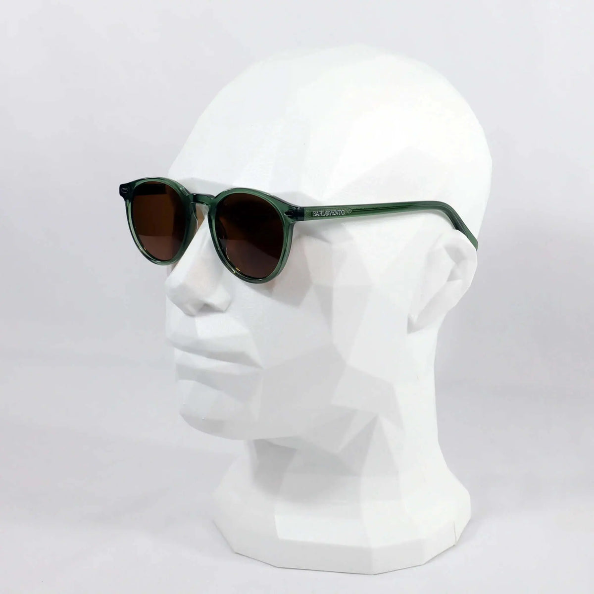 Gafas de sol redondas con montura de acetato verde translúcido y lentes oscuras marrones sobre maniquí blanco.