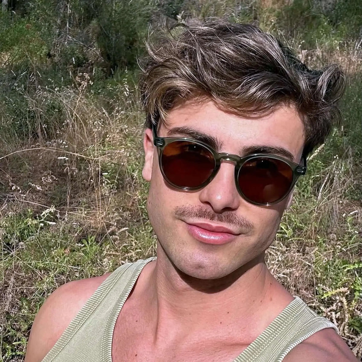 Joven con gafas de sol redondas de montura verde translúcida y lentes oscuras marrones al aire libre.