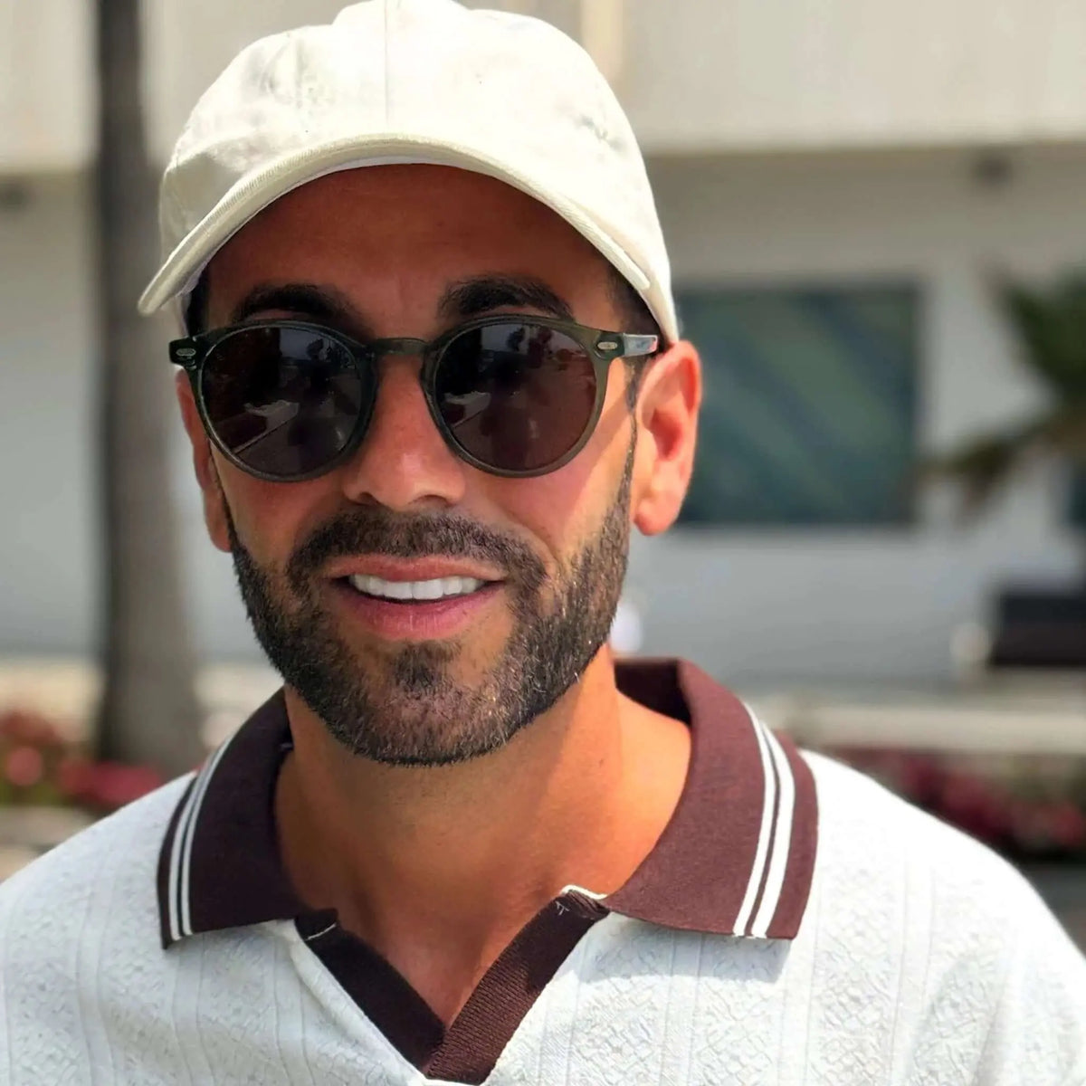 Hombre con gorra blanca y polo, usando gafas de sol redondas de montura verde y lentes marrones.