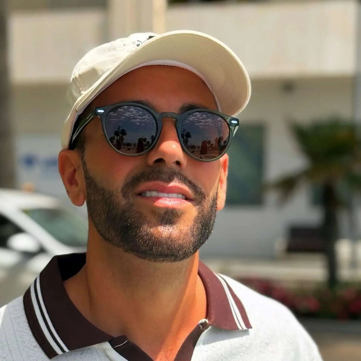 Hombre con gafas de sol redondas de montura verde oscura y lentes oscuras, usando gorra y polo casual.