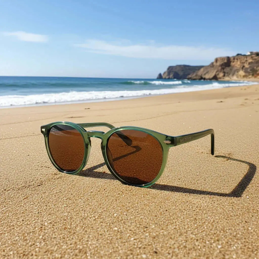 Gafas de sol redondas con montura verde translúcida y lentes marrones sobre la arena de una playa.