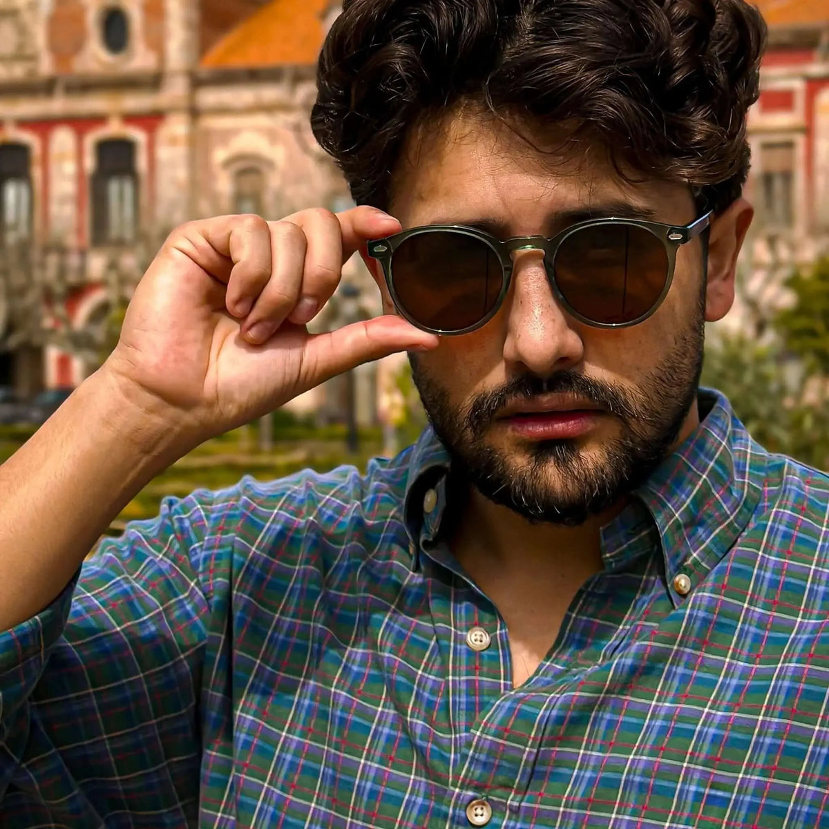 Hombre con gafas de sol redondas de montura verde translúcida y lentes marrones, vistiendo camisa de cuadros.