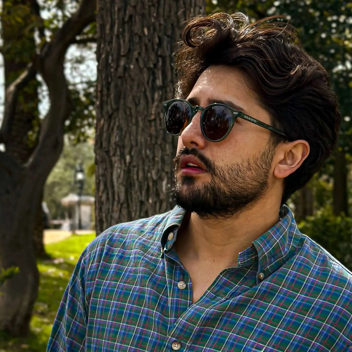 Gafas de sol de montura redonda verde y lentes marrones sobre un hombre con camisa de cuadros en un parque.