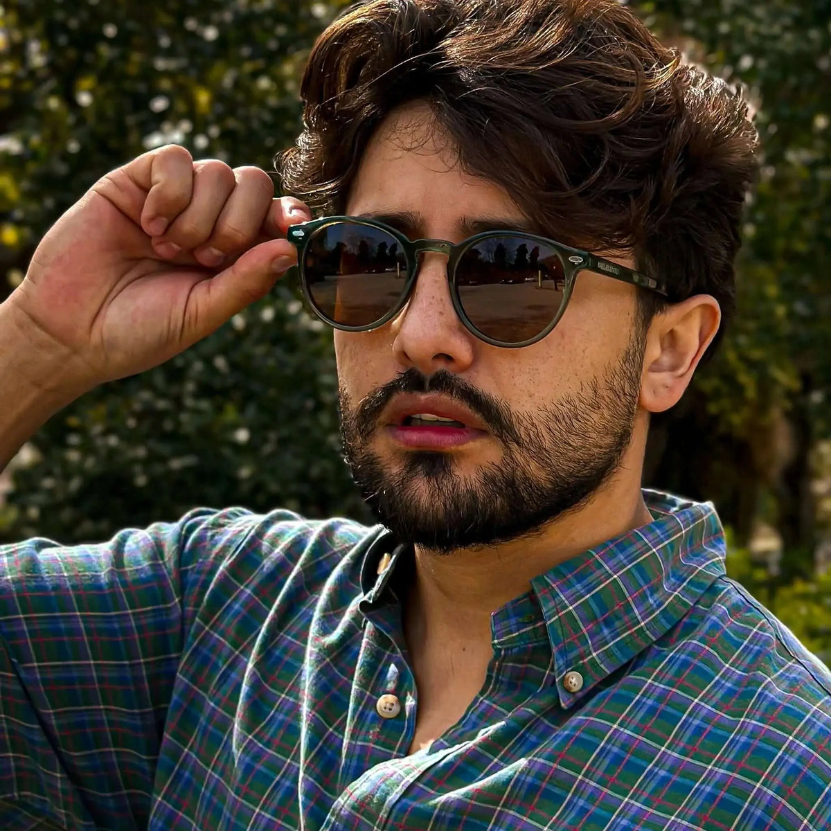 Gafas de sol redondas con montura verde translúcida y lentes marrones oscuras, usadas por un hombre con camisa a cuadros.
