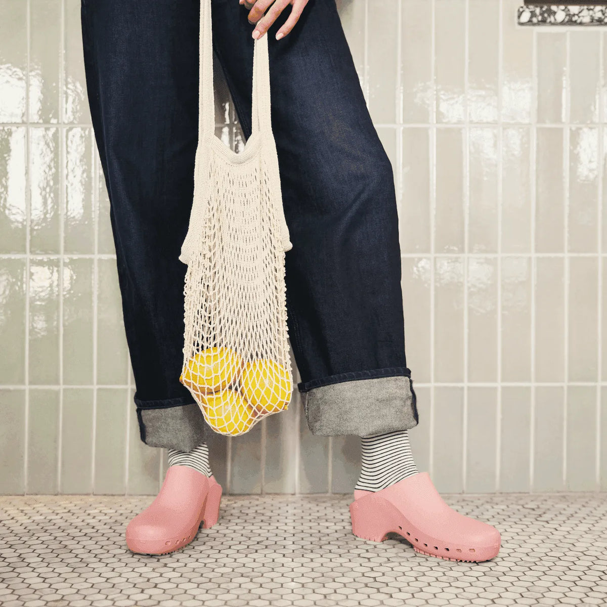 Pink clogs with perforations, paired with denim jeans and a netted tote bag, exuding a casual, modern style.