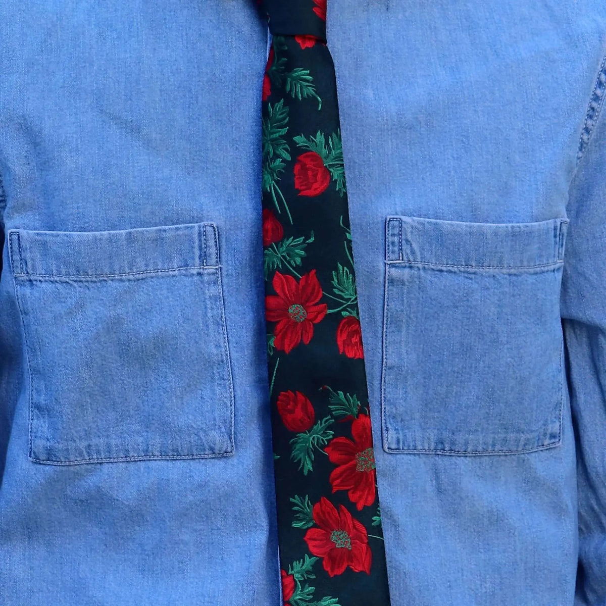Corbata negra con estampado de flores rojas y hojas verdes sobre camisa de mezclilla azul con bolsillos.
