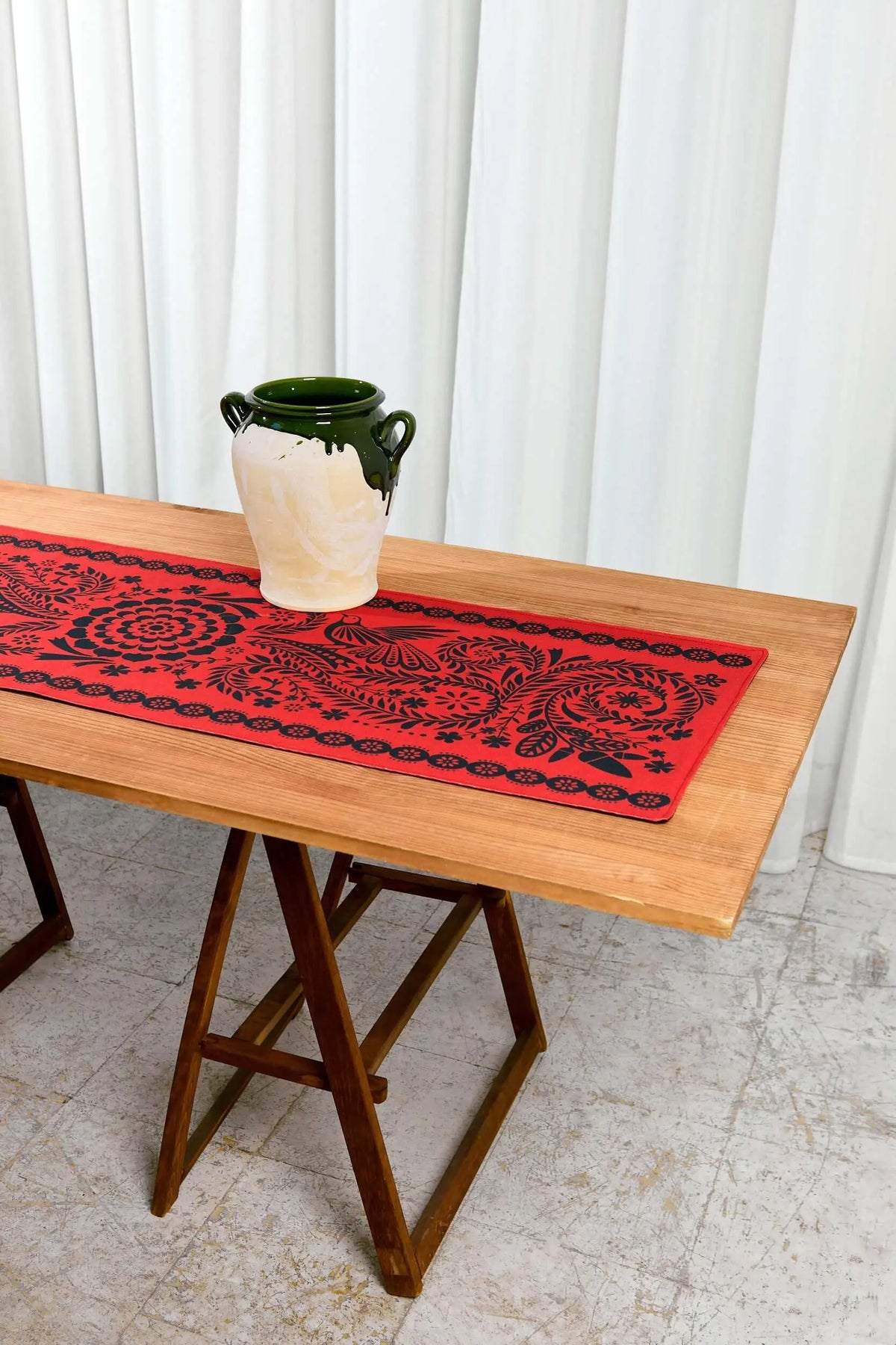 Camino de mesa rojo con estampado floral y de aves en negro, estilo folklore sobre mesa de madera rústica.