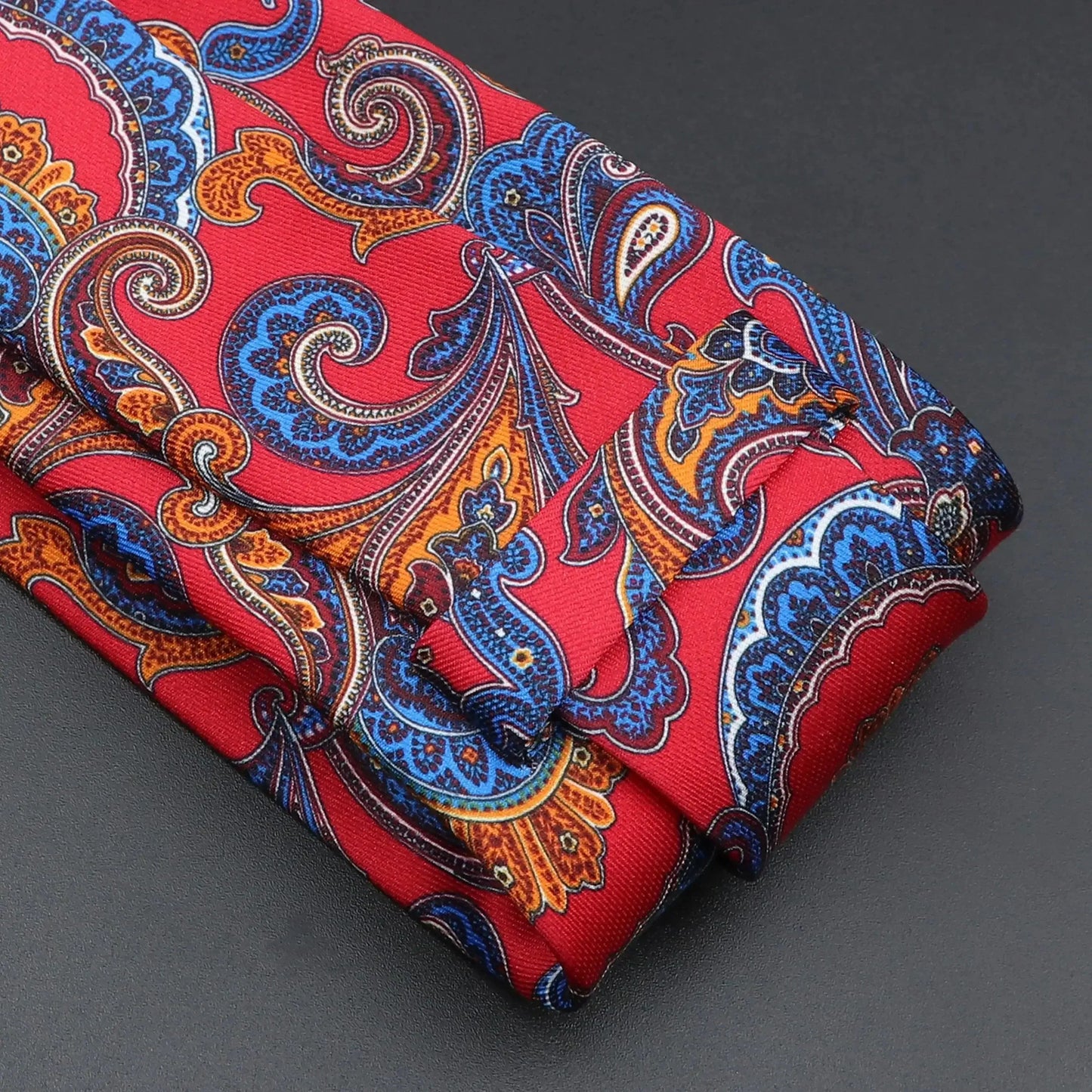 Corbata roja con estampado paisley detallado en tonos azul y naranja, sobre fondo de tela texturizada.
