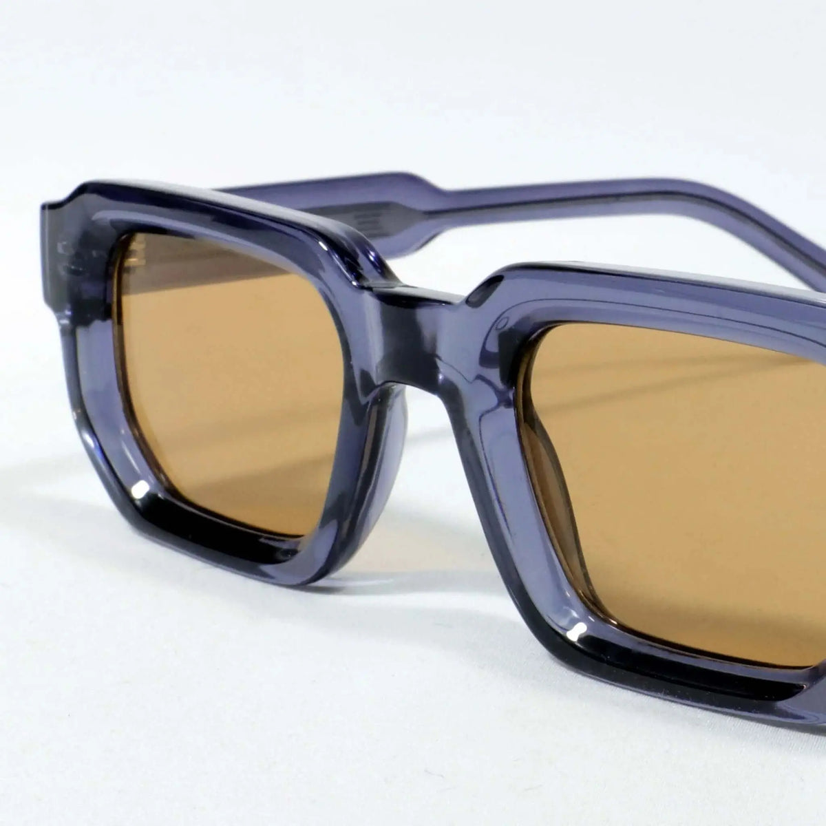 Gafas de sol de montura gruesa azul traslúcido y lentes cuadradas de color marrón, sobre fondo blanco.