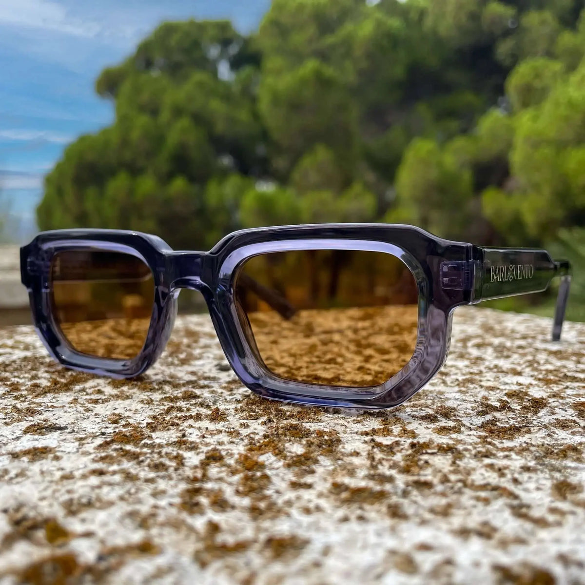 Gafas de sol rectangulares con montura de acetato azul translúcido y lentes marrones sobre una superficie de piedra.