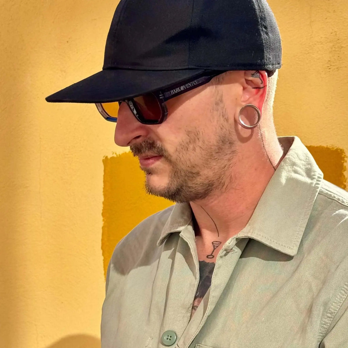 Gafas de sol de montura azul y lentes marrones en perfil de hombre con gorra negra sobre fondo amarillo.