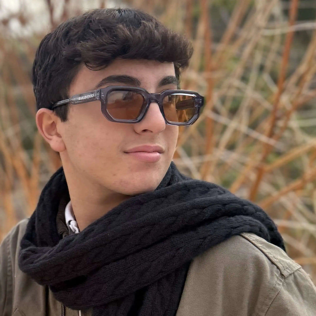 Gafas de sol rectangulares con montura azul traslúcida y cristales marrones puestas en un joven con bufanda negra.