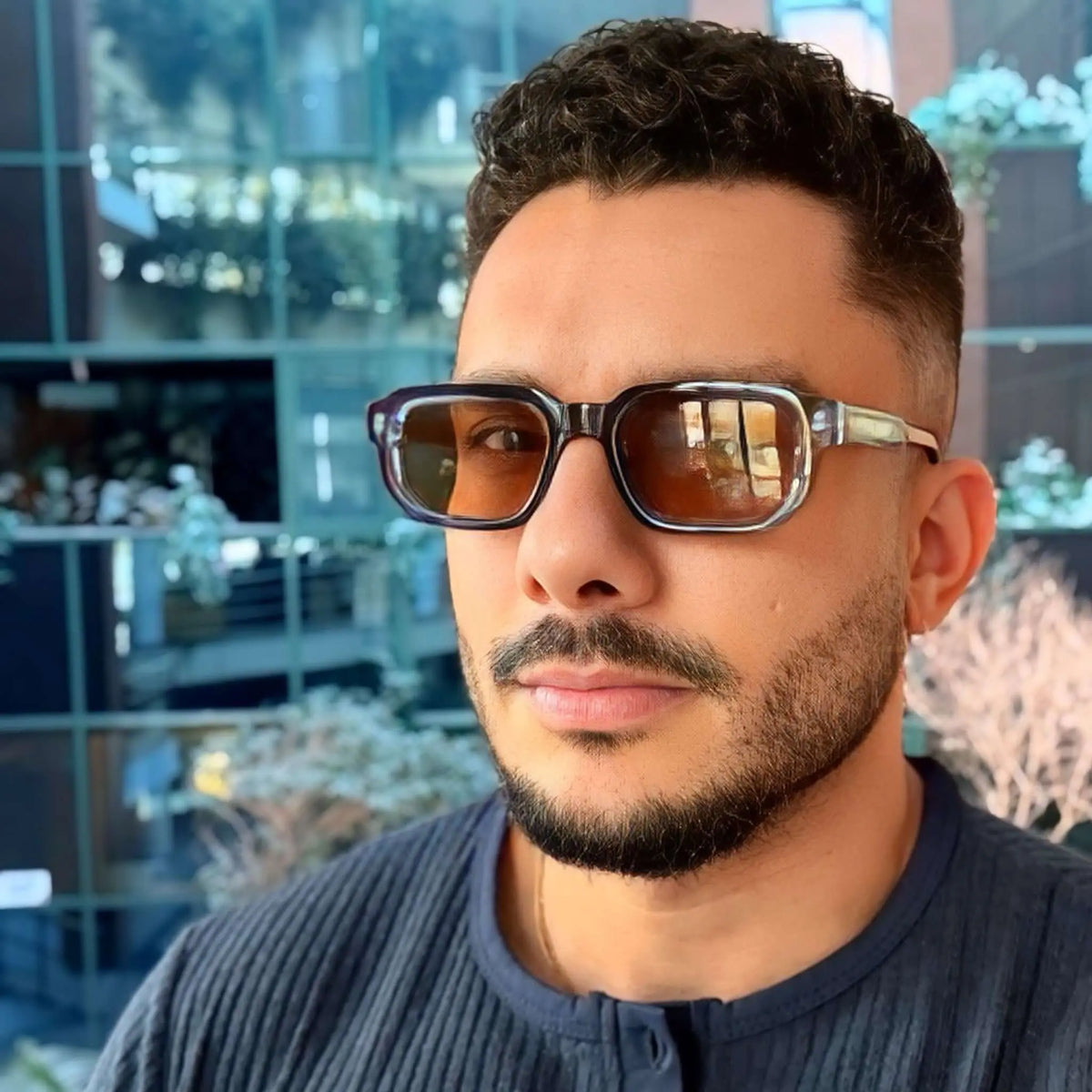 Gafas de sol de montura rectangular azul translúcida y lentes marrones, puestas en un hombre con barba.