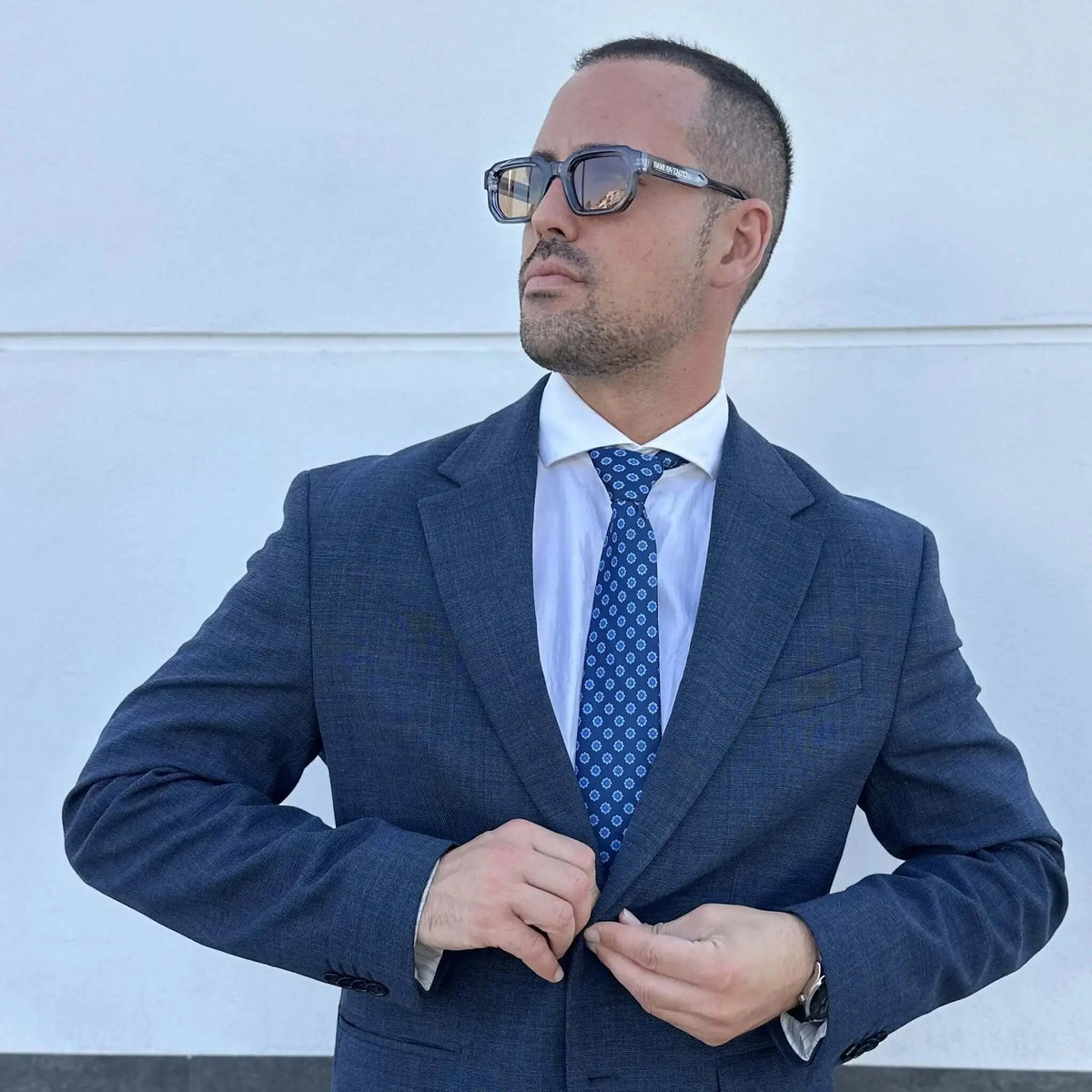 Hombre con traje azul y corbata estampada usando gafas de sol rectangulares de montura azul y lentes marrones.