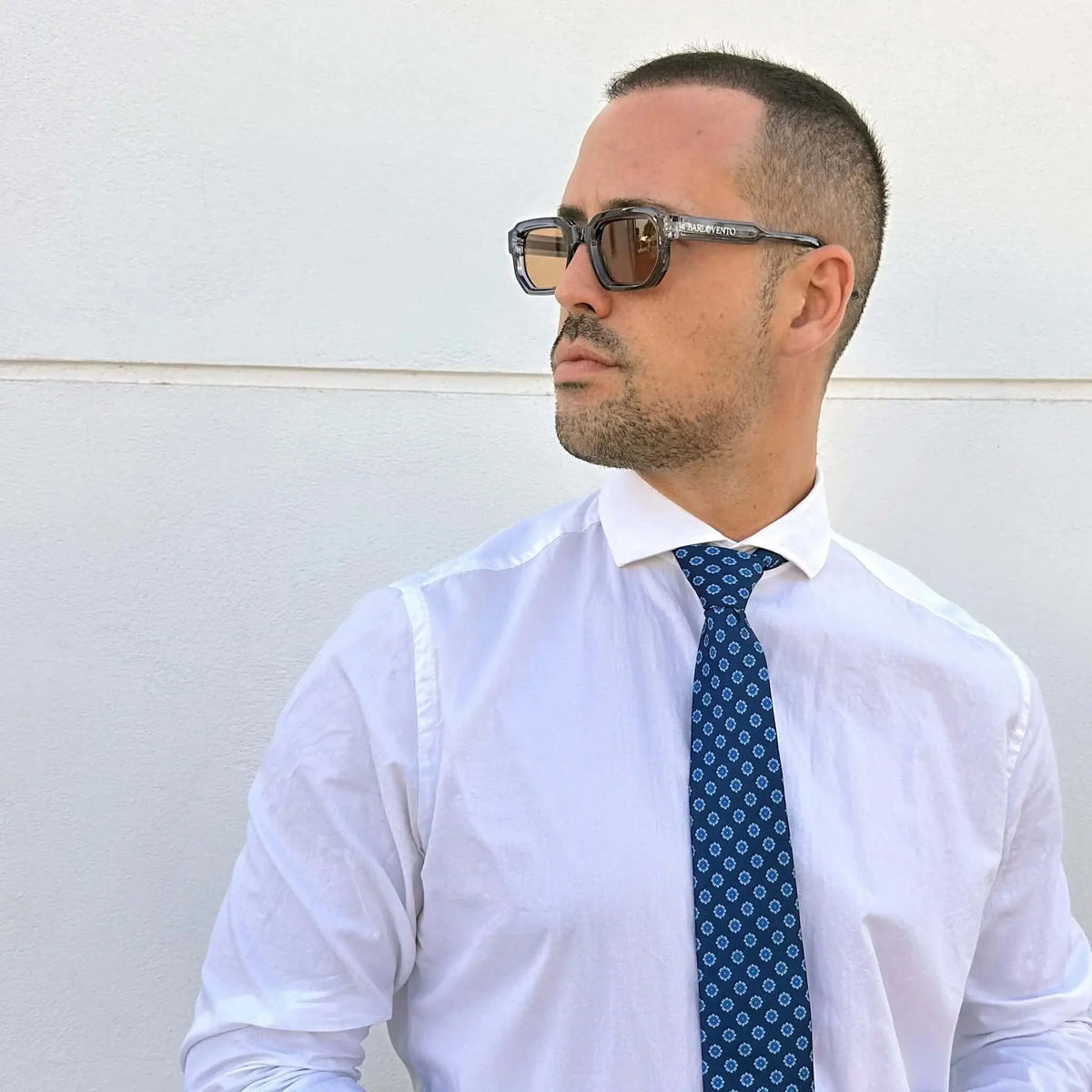 Hombre con gafas de sol de montura translúcida azulada y lentes marrones cuadradas, camisa blanca y corbata estampada.