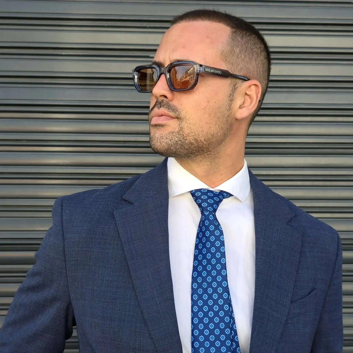 Hombre con traje azul y gafas de sol rectangulares con montura gris traslúcida y lentes color café.
