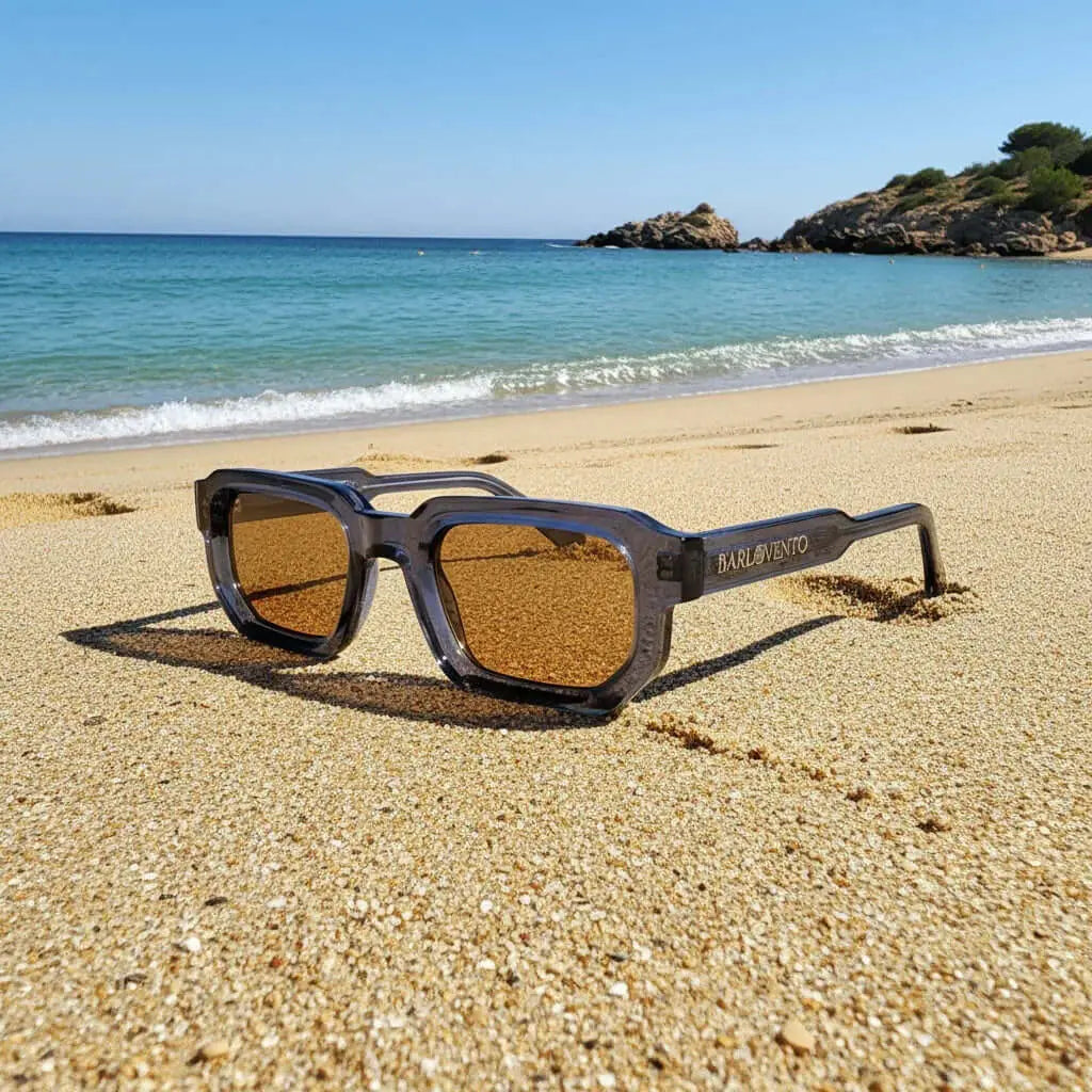 Gafas de sol rectangulares con montura azul translúcida y lentes marrones sobre la arena de una playa.