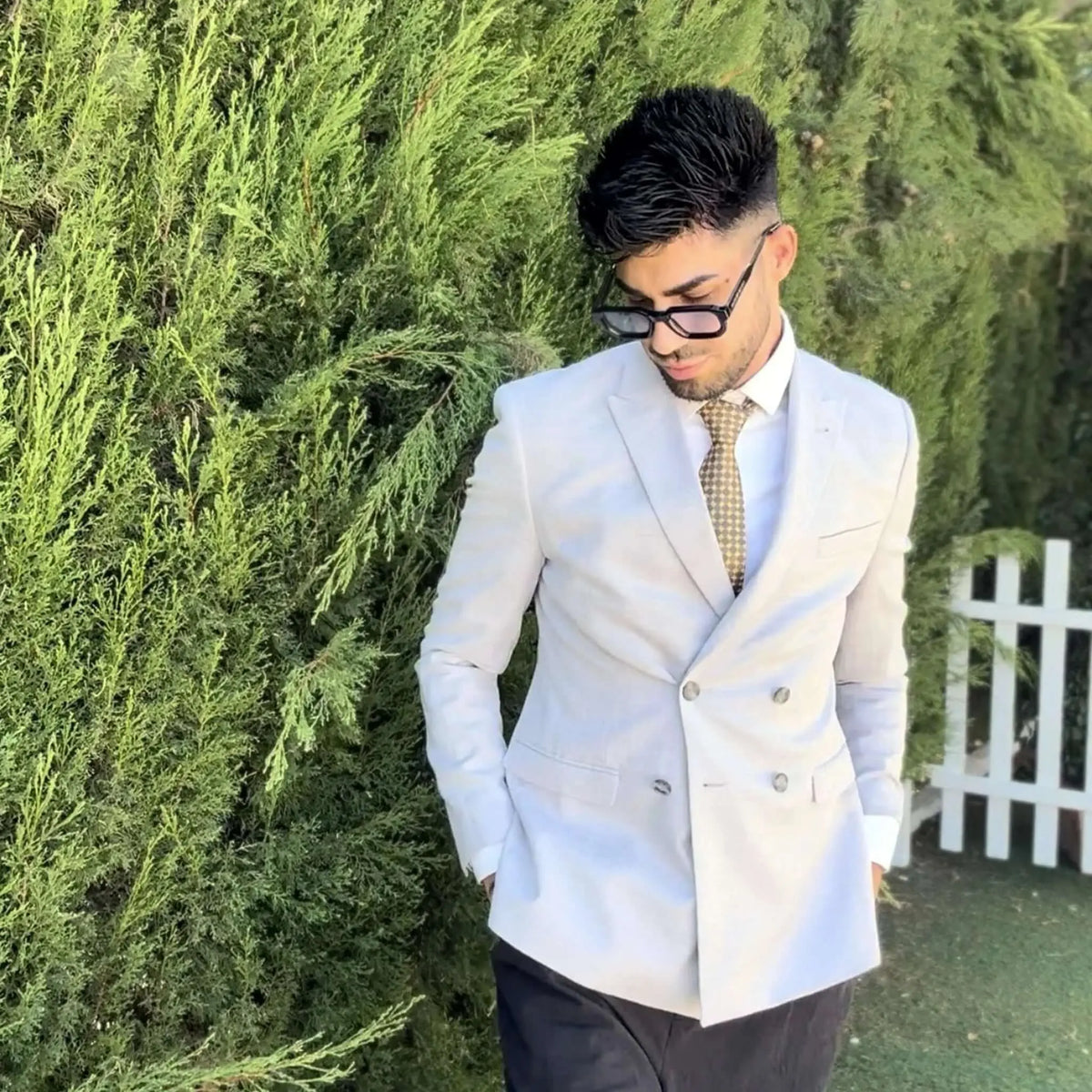 Hombre con gafas de sol de montura negra y lentes azuladas, luciendo un traje claro cruzado con corbata estampada.