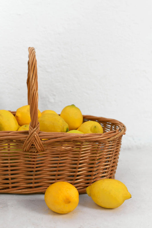 Oval natural wicker basket with a tall handle, filled with bright yellow lemons on a white textured background.