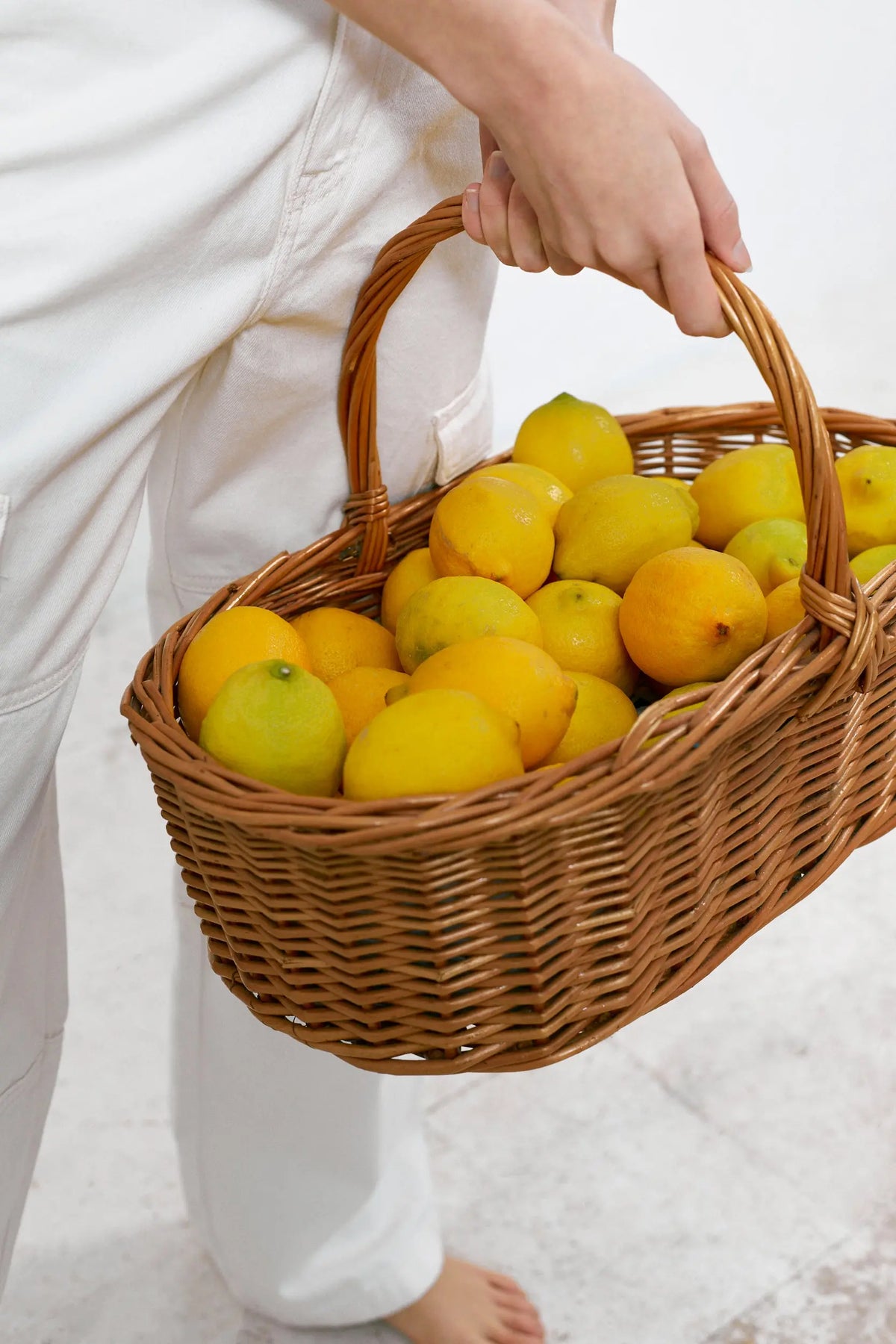 Cesta ovalada de mimbre natural con asa, llena de limones frescos, sostenida por una persona con ropa blanca.