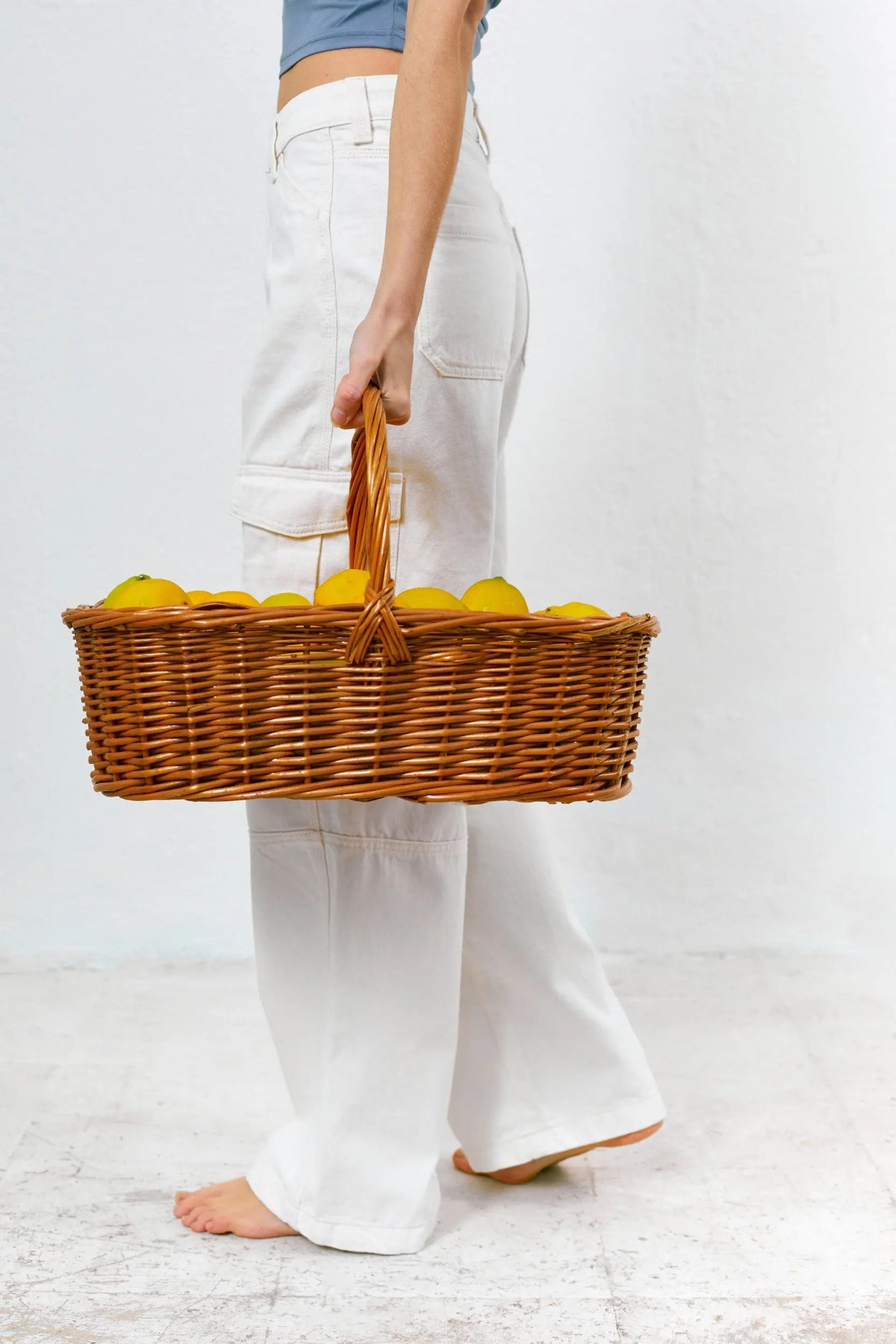Oval natural wicker basket with a handle, filled with fresh yellow lemons, held by a person in white trousers.